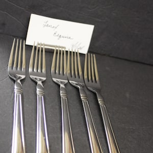 May include: Six silver forks with a classic design are arranged on a dark surface. The forks have a simple, elegant handle and tines. A small white card with handwritten text is in the background. The forks are likely for a formal dining setting.