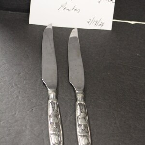 May include: Two silver knives with ornate handles featuring a design of a person in a boat. The knives are labeled "Hardanger Hodlestone Pewter 2/19/24".