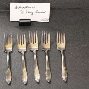May include: Five silver forks with ornate handles. The forks are arranged in a row on a dark gray surface. The text "International De Sency Rosalind 1/01/05" is written on a white card above the forks.