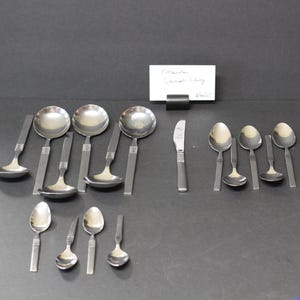 May include: A set of 12 stainless steel spoons, including serving spoons and teaspoons. The spoons are arranged on a gray surface.