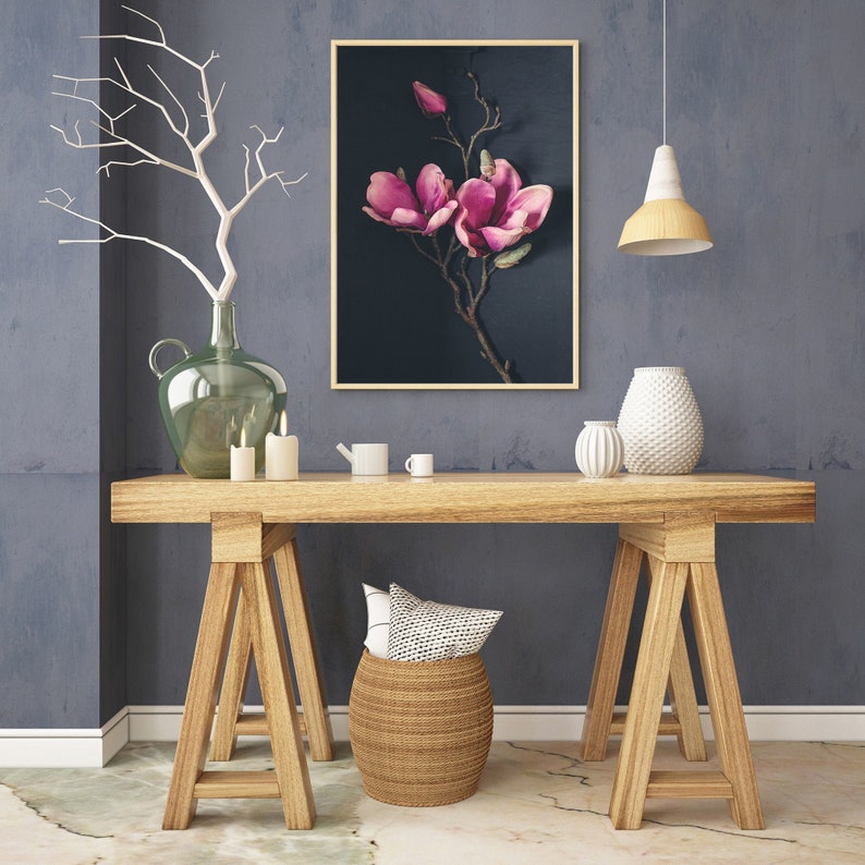 May include: A wooden table with two trestle legs, a wicker basket, a green glass vase, and a framed print of pink magnolia flowers on a dark background.
