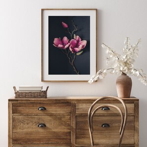 May include: A framed print of a pink magnolia flower branch against a dark blue background. The print is hanging on a white wall above a wooden dresser with drawers and a wooden chair.