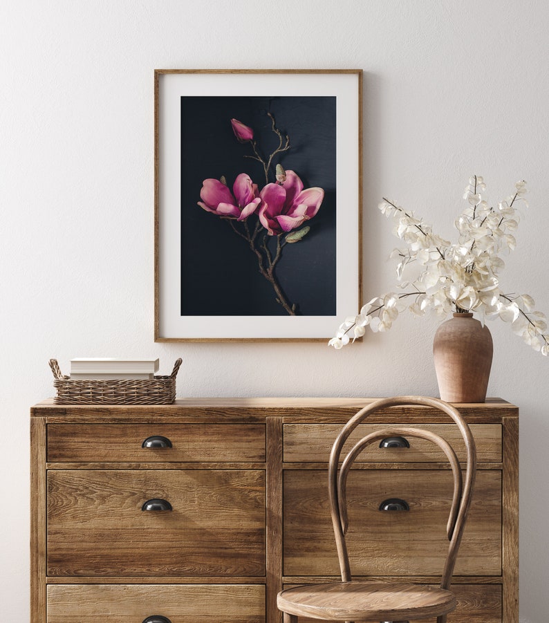 May include: A framed print of a pink magnolia flower branch against a dark blue background. The print is hanging on a white wall above a wooden dresser with drawers and a wooden chair.