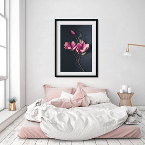 May include: A framed print of a pink magnolia flower branch against a dark blue background. The print is hanging on a white wall above a bed with pink and white bedding.