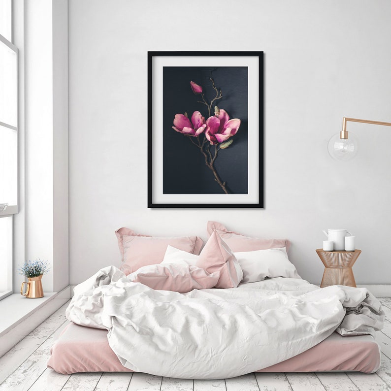 May include: A framed print of a pink magnolia flower branch against a dark blue background. The print is hanging on a white wall above a bed with pink and white bedding.