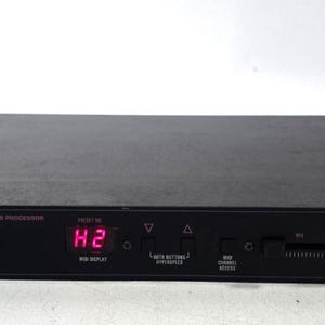 Puede incluir: Un procesador de efectos digitales ART MultiVerb LT negro con chasis para montaje en rack. El panel frontal cuenta con una pantalla LED roja que muestra H2, varios botones de control, un deslizador y texto en rosa. Un cable de alimentación negro está conectado.