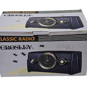 Puede incluir: Una caja de producto de cartón blanco para una radio Crosley Classic Radio. La caja presenta texto en negro y amarillo, rayas negras horizontales y una ilustración gráfica de una radio azul oscuro con un dial circular grande y dos perillas.