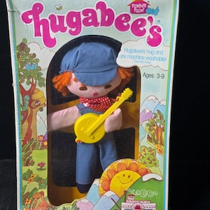 Può includere: Una bambola vintage Huggabee's di nome Huck, vestita con una salopette blu e un berretto blu, che tiene un banjo giallo. La bambola è lavabile in lavatrice ed è adatta per bambini dai 3 ai 9 anni. La scatola presenta un'illustrazione colorata di un sole e una barca.