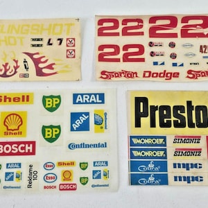 Puede incluir: Una colección de calcomanías de automóviles antiguos. Incluye calcomanías con el texto "SLINGSHOT", "22", "426 CU. IN.", y logotipos de marcas como "Esso", "Shell", "BP", "Aral", "Bosch" y "Prestone".