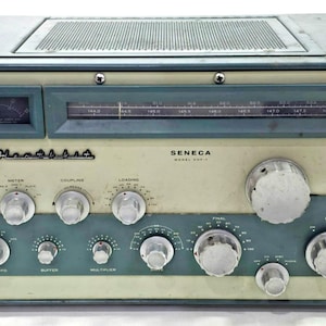 Puede incluir: Un transmisor de radio vintage Heathkit Seneca modelo VHF-1 en una carcasa de metal azul y blanco roto. El panel frontal presenta una pantalla de frecuencia analógica rectangular, un pequeño medidor, numerosos mandos plateados y un botón rojo.