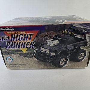 Op de afbeelding: Doos van een op afstand bestuurbare RadioShack 4x4 Night Runner off-road voertuig. De zwarte truck heeft grote banden, een rolkooi en de tekst "Night Runner" op de zijkant. De doos toont de truck en de tekst "RadioShack".