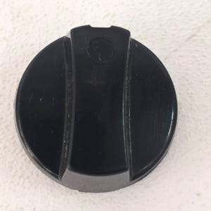 May include: A black, circular control knob with a slightly raised center section. The knob has a smooth, glossy finish and a small, circular indentation near the top. The design features vertical grooves on either side of the center.