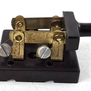 May include: A vintage electrical switch featuring brass components and a black base. The switch has a lever and two silver screws. The brass parts show signs of age. Designed for electrical circuits, it shows signs of wear.