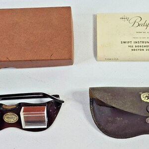 May include: Vintage Swift Bedspecks, including a brown leather case, a cardboard box, and an instruction booklet. The Bedspecks feature a black frame and prism lenses. The case and box are brown. The instruction booklet has the text "Swift Instruments, Inc."