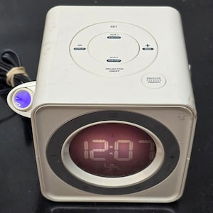 Puede incluir: Radio despertador digital blanco con una pantalla digital roja que muestra las 12:07. El reloj tiene botones para configurar la hora, la alarma y la proyección. El reloj también tiene un botón de repetición y un botón de atenuación.