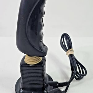 May include: Black joystick with a red button on top. The joystick has a curved handle with finger grooves and a beige spring base. A black cable is attached, secured with a rubber band, and ends in a connector.