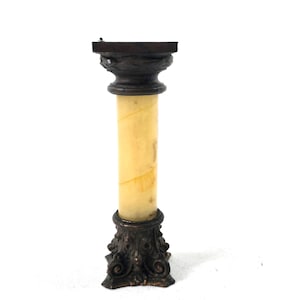 May include: A small, decorative column with a square top and ornate base. The column has a light yellow, cylindrical body with brown accents at the top and bottom. The base and top are dark brown with detailed, swirling designs. The column stands approximately 6 inches tall.
