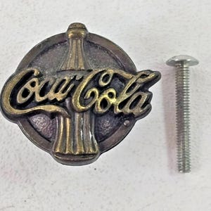 May include: A vintage-style Coca-Cola emblem with a screw. The emblem features the Coca-Cola logo in gold lettering over a bottle outline, set against a dark, circular background. The screw is silver and has a rounded head.