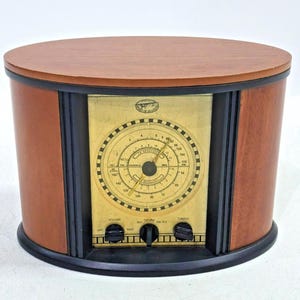May include: An oval-shaped radio with a wooden exterior and a central gold-colored faceplate. The faceplate features a circular analog tuning dial with black markings and numbers, three black control knobs, and the text Spirit of St. Louis.