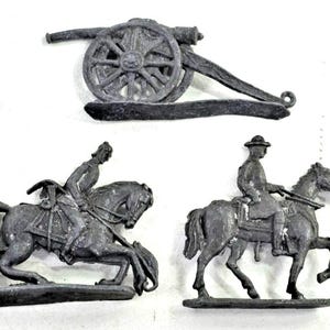 May include: A set of five grey plastic toy figures including two standing soldiers aiming rifles, two soldiers mounted on horses, and one artillery cannon on wheels. Each figure is moulded in a single colour with detailed military uniforms and gear.