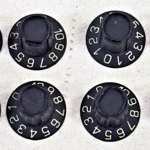 May include: Eight black circular control knobs arranged in two rows. Each knob features a fluted side and a flat top surface printed with white numbers. Four knobs display a scale from 1 to 10, while the other four display a scale from 0 to 5.