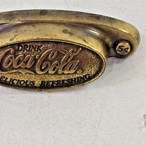 May include: Vintage brass Coca-Cola drawer pull with the text "DRINK Coca-Cola DELICIOUS REFRESHING". Includes two silver screws. The pull has a curved design and the number "5¢" is on each end.