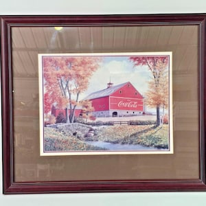 May include: Framed print featuring a red barn with the Coca-Cola logo, set in a picturesque autumn landscape. The artwork includes a small stone bridge, trees with fall foliage, and a serene stream. The frame is a dark, rich brown.