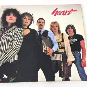 May include: Album cover for the band Heart. The cover features five people, with the band's name in red script. The person on the left wears a black and white striped jacket. The person on the right wears a black t-shirt and blue jeans.