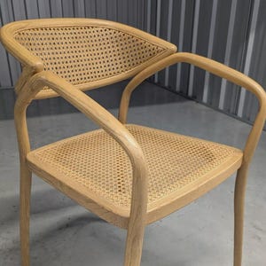 May include: Light wood-toned chair with a woven cane seat and backrest. The chair features curved armrests and legs, with a natural finish. The backrest has a unique, slightly curved shape. The chair is designed for comfort and style.