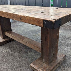 May include: A rustic wooden table with a rectangular top and sturdy, square legs. The table is made of reclaimed wood with visible grain and knots. Metal corner brackets reinforce the edges. The table is approximately 183 cm long, 91 cm wide, and 76 cm high.