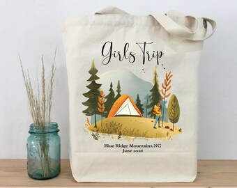 May include: Cream-colored tote bag with a camping scene illustration. The design features an orange tent, trees, and a person. Text reads "Girls Trip" and "Blue Ridge Mountains, NC June 2026".