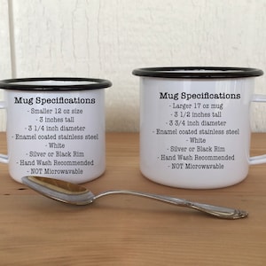 May include: Two white enamel mugs with black rims and handles, one smaller (7.6 cm tall, 8.3 cm diameter) and one larger (8.9 cm tall, 9.5 cm diameter). Text on the mugs details specifications. A silver spoon rests in front.