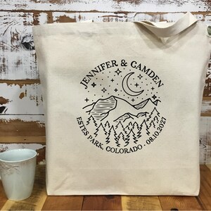 以下が含まれることがあります： 黒い山と月のデザインが施されたクリーム色のトートバッグ。「JENNIFER & CAMDEN ESTES PARK, COLORADO 08.10.2021」と書かれています。左下には水色のマグカップが見えます。