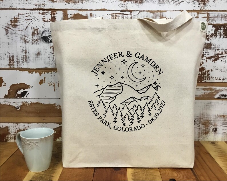 以下が含まれることがあります： 黒い山と月のデザインが施されたクリーム色のトートバッグ。「JENNIFER & CAMDEN ESTES PARK, COLORADO 08.10.2021」と書かれています。左下には水色のマグカップが見えます。