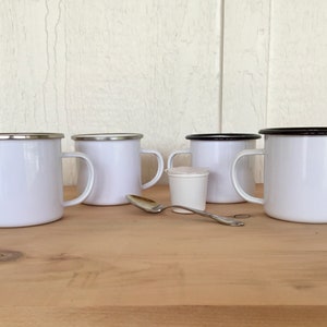 May include: Four white enamel mugs with silver and black rims are arranged on a wooden surface. A small white container and a silver spoon are also present. The mugs have a classic design, suitable for camping or everyday use.