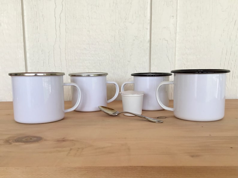May include: Four white enamel mugs with silver and black rims are arranged on a wooden surface. A small white container and a silver spoon are also present. The mugs have a classic design, suitable for camping or everyday use.