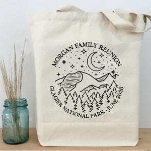 以下が含まれることがあります： 黒いグラフィックデザインのクリーム色のトートバッグ。デザインは山脈、木、星、三日月を特徴とし、「MORGAN FAMILY REUNION」と「GLACIER NATIONAL PARK JUNE 2026」の文字が書かれています。
