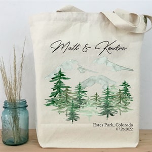 May include: A white canvas tote bag with a watercolor illustration of a mountain range and pine trees. The text "Matt & Kendra" is printed in black cursive script at the top of the bag. The text "Estes Park, Colorado 07.26.2022" is printed in black at the bottom of the bag.