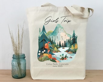 Könnte beinhalten: Eine weiße Canvas-Tragetasche mit einer bunten Illustration einer Bergszene und einer Person beim Wandern. Der Text "Girls Trip" ist oben auf der Tasche in Kursivschrift geschrieben. Der Text "Estes Park, Colorado June 2025" ist unter der Illustration gedruckt.