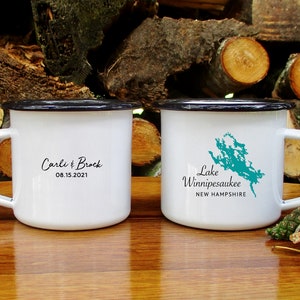 May include: Two white enamel mugs with black rims. One mug has the text "Carli & Brock 08.15.2021" and the other mug has the text "Lake Winnipesaukee NEW HAMPSHIRE" and a teal outline of the lake.