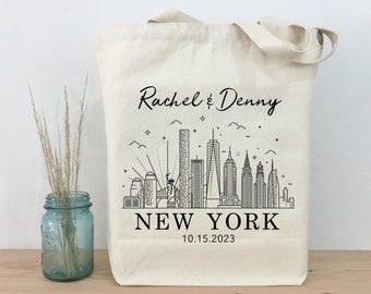 May include: A white canvas tote bag with black text and a black line drawing of the New York City skyline. The text reads "Rachel & Denny" and "NEW YORK" with the date "10.15.2023" below the city skyline.