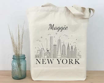 Könnte beinhalten: Eine weiße Canvas-Tragetasche mit einer schwarzen Umrandung der Skyline von New York City und dem aufgedruckten Text "NEW YORK" auf der Vorderseite. Der Name "Maggie" ist über der Skyline aufgedruckt.