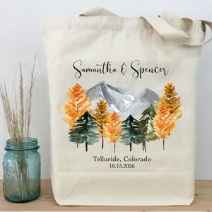 May include: Cream-colored tote bag featuring a watercolor design of a mountain range and trees in shades of orange and green. The names "Samantha & Spencer" and the location "Telluride, Colorado 10.15.2026" are printed on the bag.