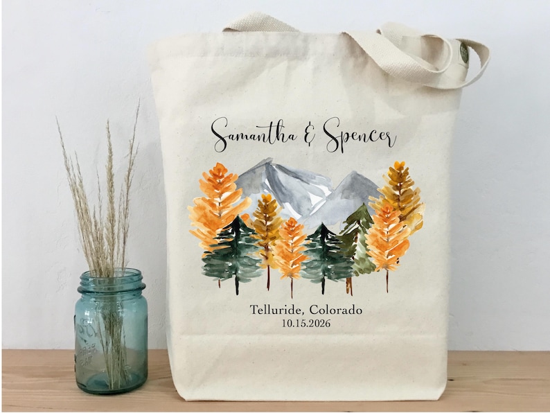 May include: Cream-colored tote bag featuring a watercolor design of a mountain range and trees in shades of orange and green. The names "Samantha & Spencer" and the location "Telluride, Colorado 10.15.2026" are printed on the bag.