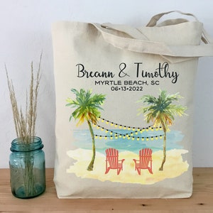 May include: A white canvas tote bag with a beach scene design. The bag has black text that reads "Breann & Timothy Myrtle Beach, SC 06-13-2022". The design features two palm trees, a beach, and two red beach chairs.