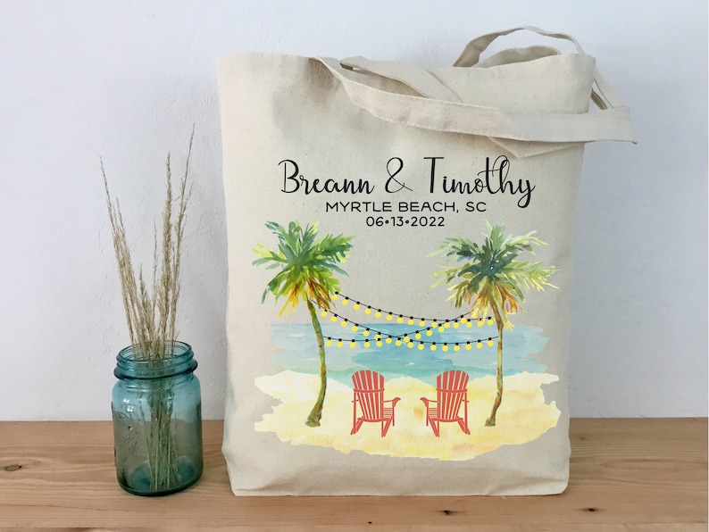 May include: A white canvas tote bag with a beach scene design. The bag has black text that reads "Breann & Timothy Myrtle Beach, SC 06-13-2022". The design features two palm trees, a beach, and two red beach chairs.