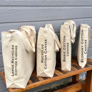 May include: Four beige canvas tote bags of varying sizes are displayed on a wooden bench. The bags have long handles and black text that reads "Large Recycled Cotton Canvas", "Recycled Cotton Canvas", "Basic Cotton Canvas", and "Economy Cotton Canvas".