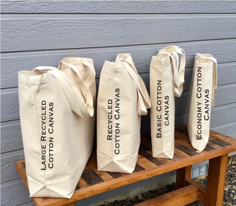 May include: Four beige canvas tote bags of varying sizes are displayed on a wooden bench. The bags have long handles and black text that reads "Large Recycled Cotton Canvas", "Recycled Cotton Canvas", "Basic Cotton Canvas", and "Economy Cotton Canvas".