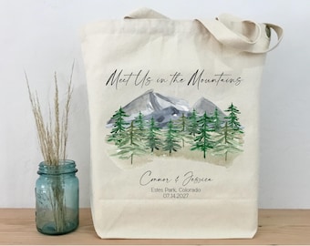 Könnte beinhalten: Cremefarbene Tragetasche mit einem Aquarell-Design von Bergen und Wald. Der Text "Meet Us in the Mountains" befindet sich über der Illustration. Darunter stehen die Namen "Connor & Jessica" und das Datum "07.14.2027".