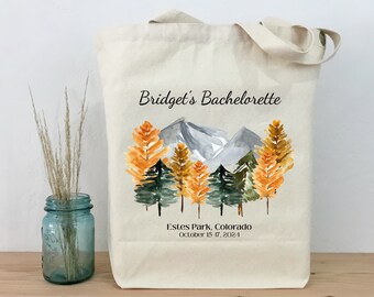 Könnte beinhalten: Eine weiße Canvas-Tragetasche mit einem Aquarell-Design von Bergen und Bäumen in Orange-, Grün- und Grautönen. Der Text "Bridget's Bachelorette" ist oben auf der Tasche gedruckt, und der Text "Estes Park, Colorado October 15-17, 2024" ist unten gedruckt.